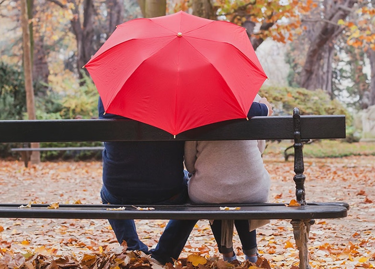 The back view of a couple sitting on a bench holding a red umbrella, surrounded by fallen leaves